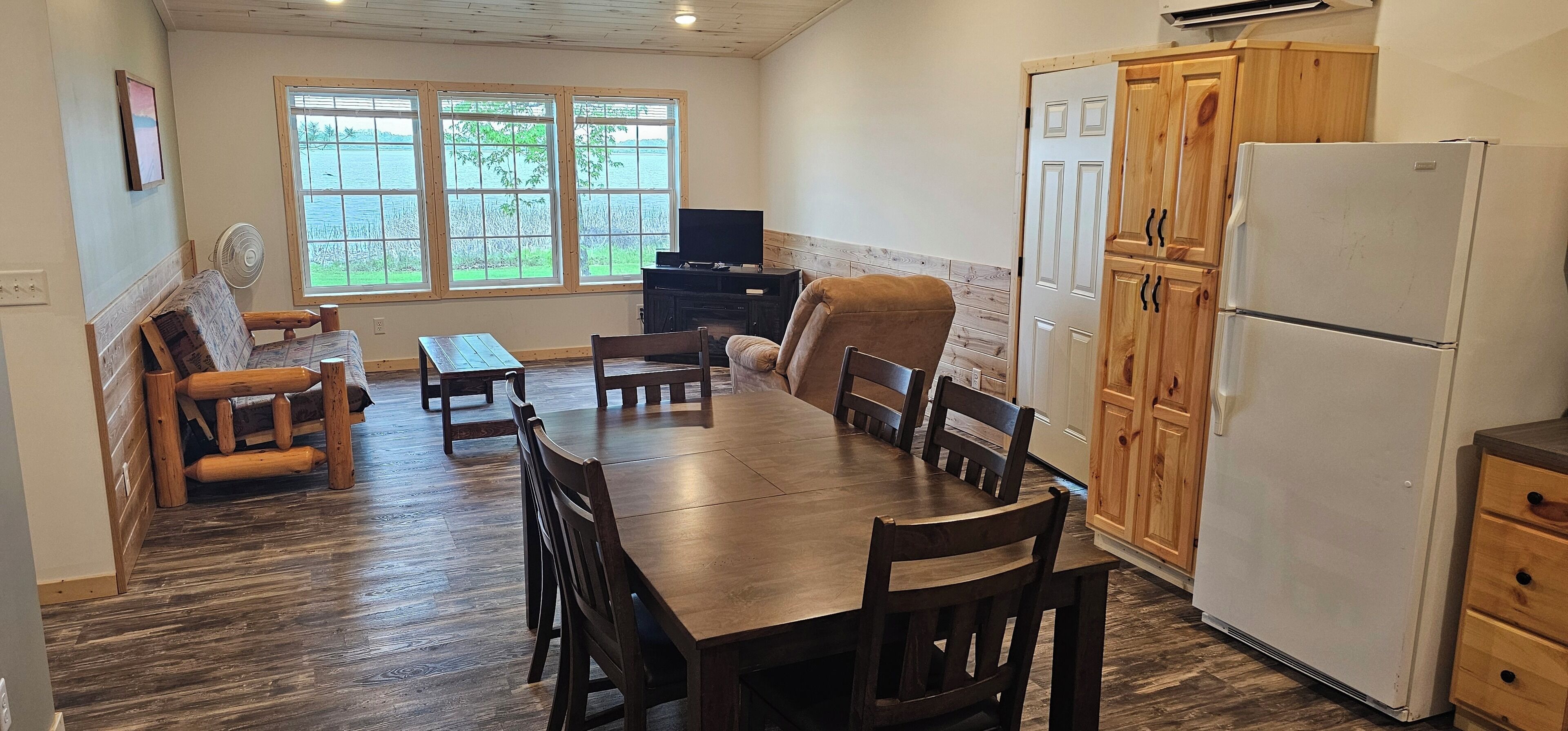 A dining area with a wooden table and chairs, a television, and a view of a body of water through large windows.