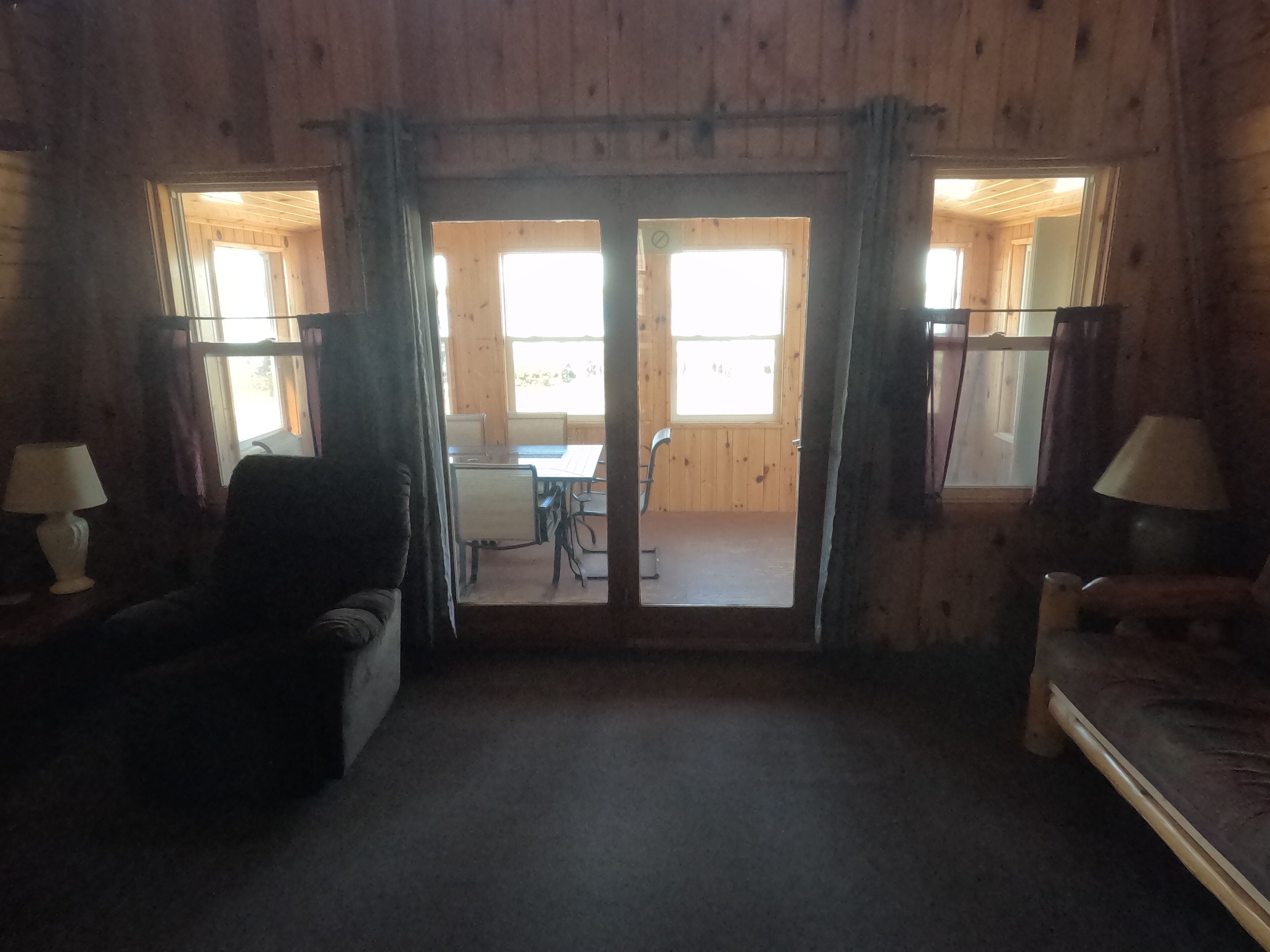 Una habitación con interior de madera, un sofá, una mesa y dos lámparas. Hay cortinas en las ventanas, y una puerta da a otra habitación.