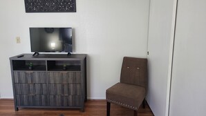 Ein Zimmer mit einem dunklen Holzschrank, einem Fernseher darauf, einer kleinen Topfpflanze und einem braunen Stuhl.