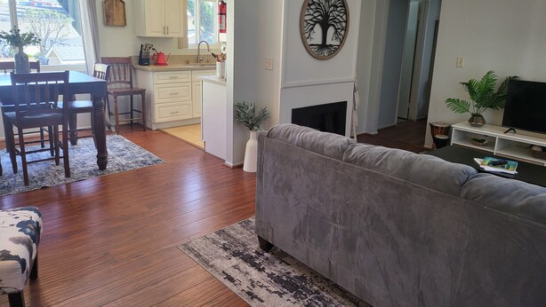 Ein Wohnzimmer mit grauem Sofa, einem Holzesstisch und Stühlen. Es gibt eine Küchenzeile mit weißen Schränken und einen Kamin. Im Wohnzimmer steht ein Fernseher auf einem Schrank.