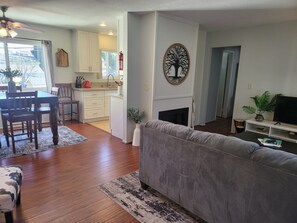 Een woonkamer met een grijze bank, een houten eettafel en stoelen. Er is een keuken met witte kasten en een open haard. Een televisie staat op een dressoir in de woonkamer.