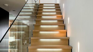 Una escalera moderna con escalones de madera y barandillas de vidrio, que conduce a un espacio con una vitrina de copas de vino.