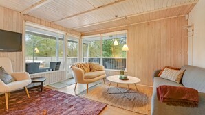 Ein gemütliches Zimmer mit einer Couch, einem Sessel und einem kleinen Tisch. Große Fenster und ein Deckenventilator sind vorhanden.
