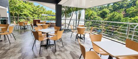 An outdoor dining area with tables and chairs, surrounded by a railing, overlooking a lush green landscape.