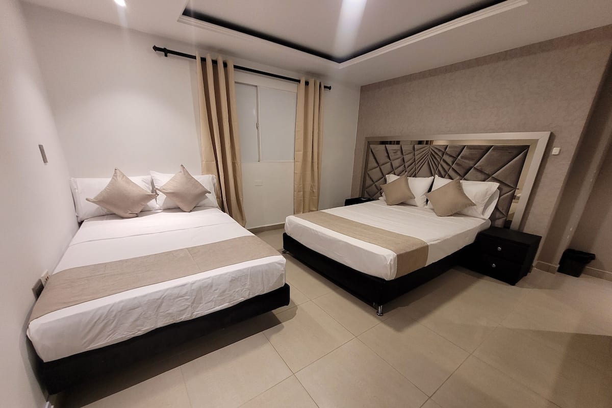 A hotel room with two beds, a large window with curtains, a modern headboard, and a bedside table.