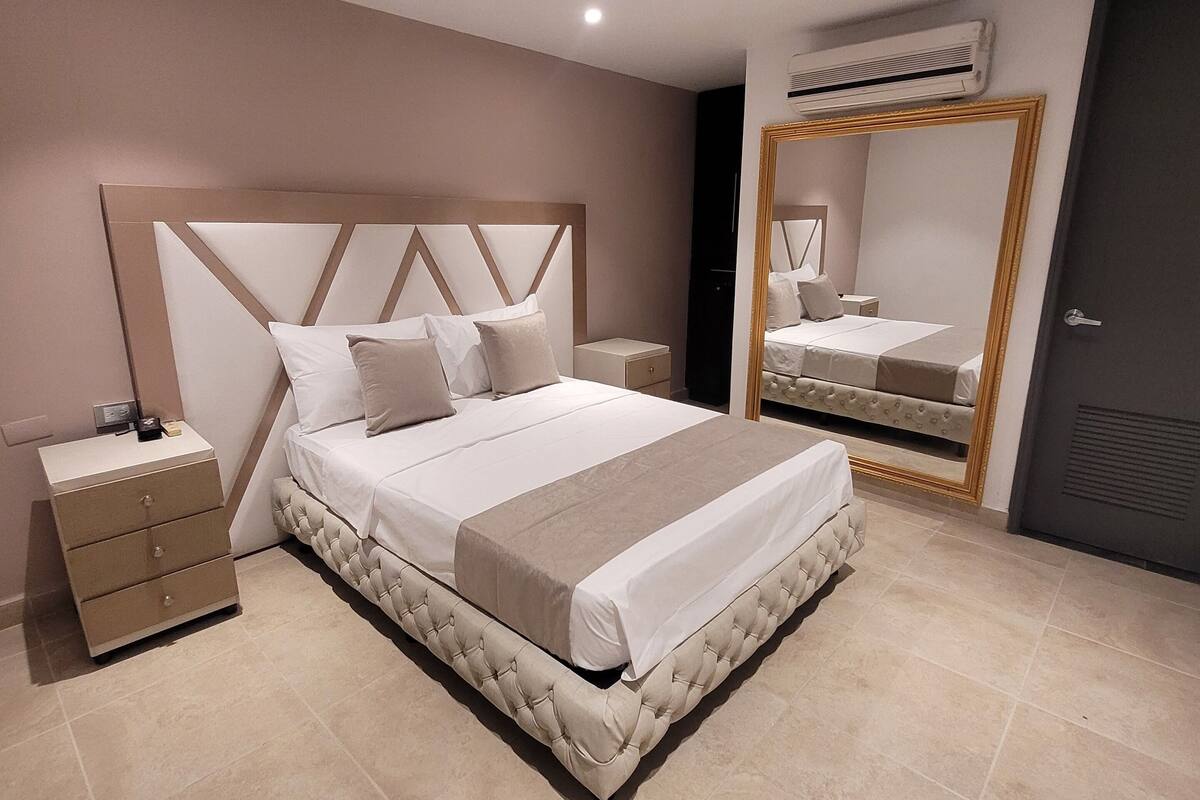 A modern bedroom with a large bed, a bedside table, a nightstand, a mirror, and an air conditioning unit.