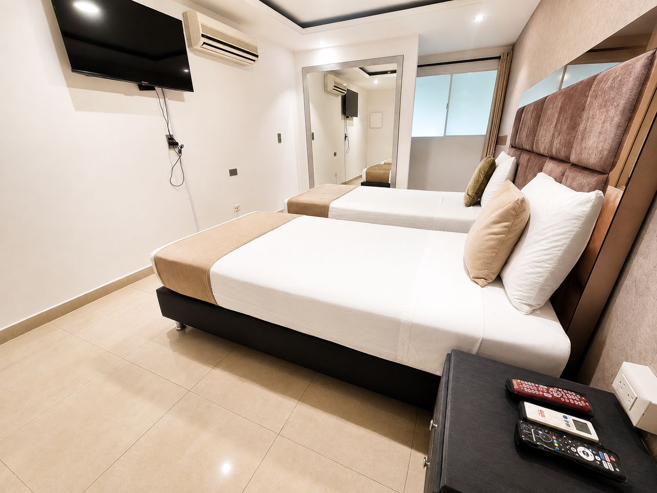 A modern hotel room with a flat-screen TV mounted on the wall, a bed with a headboard, a bedside table with a remote control, and a bathroom visible through an open door.