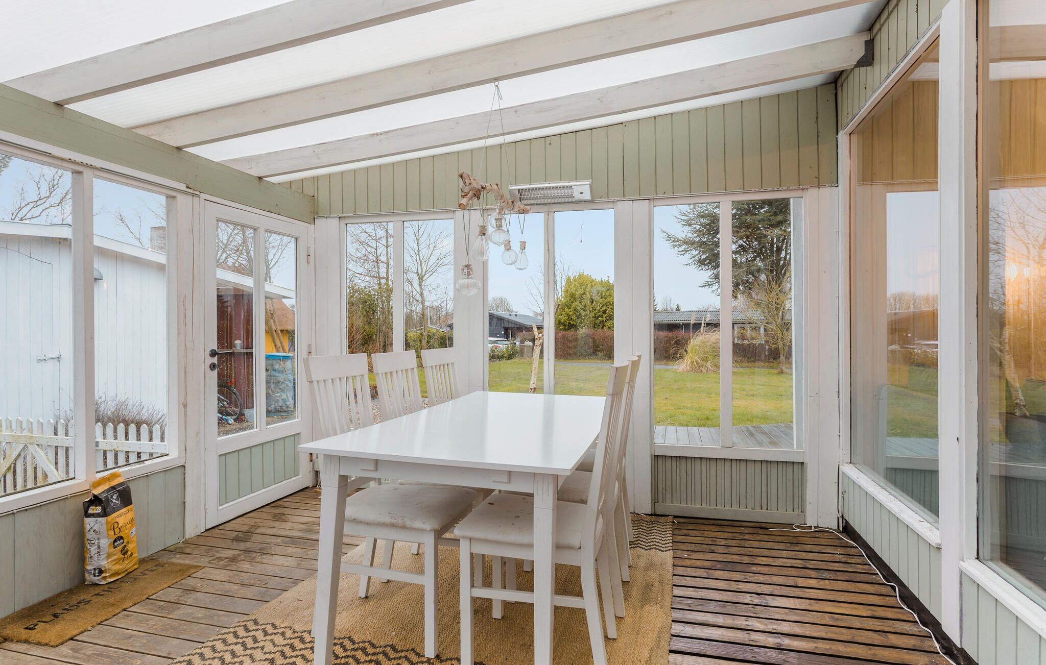 En overdækket terrasse med et hvidt bord og stole, store vinduer og udsigt til baghaven.