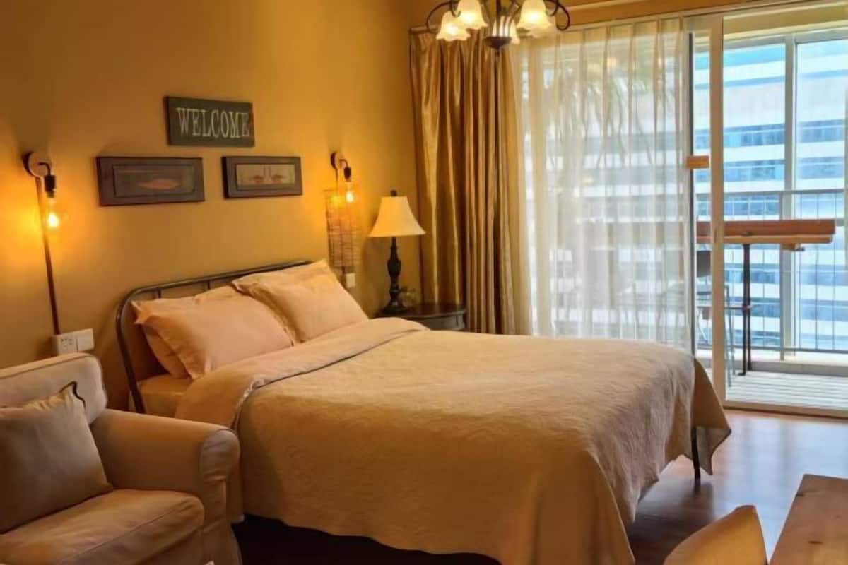 Quarto de hotel com cama, cadeira, luminária, placa 'WELCOME' e janela com cortinas.