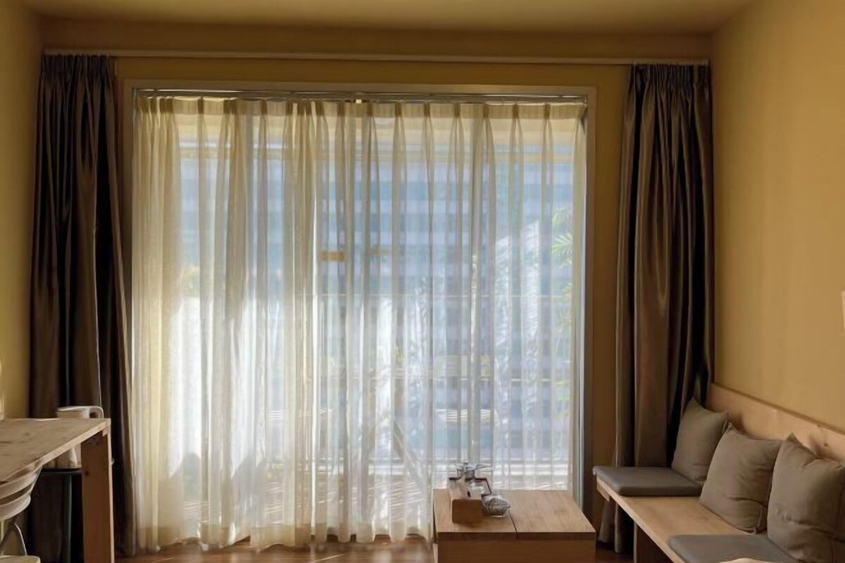 A room with a large window covered by sheer curtains, a wooden bench, and a small table with a decorative item.