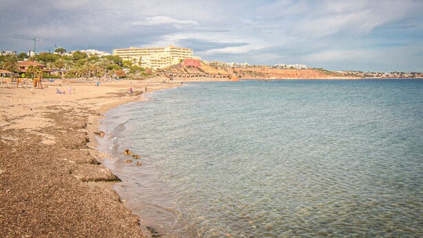 A coastal beach with a large building in the background, clear water, and a sandy shore. - 2 bedroom stunning home in La Zenia (La Zenia)