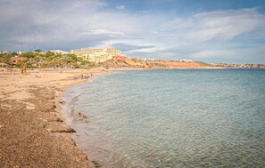 Une plage côtière avec un grand bâtiment en arrière-plan, une eau claire et un rivage sablonneux.