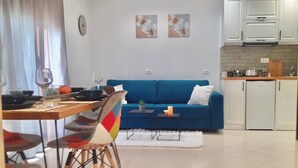 A modern living room with a blue sofa, a dining table with chairs, and a kitchen area with cabinets and a refrigerator. - Charming 1-bedroom apartment with WiFi, AC in delightful Sarandë (Sarandë)