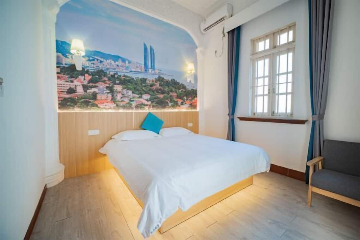 A bedroom with a bed, a window with curtains, a chair, and a cityscape mural on the wall.