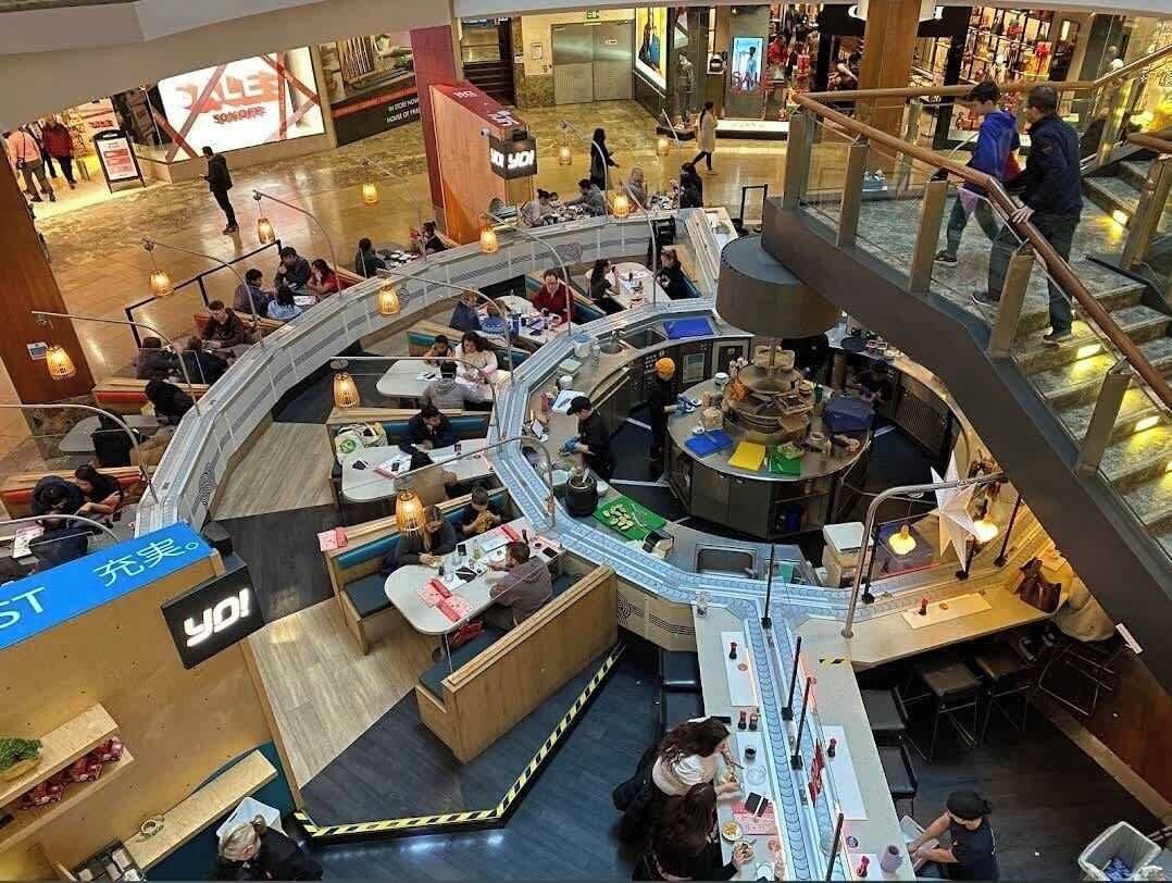 A multi-level shopping mall with a central atrium, escalators, and various retail stores.