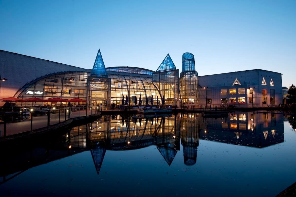 A modern building with a glass facade, a reflection in the water, and a bridge leading to the entrance.