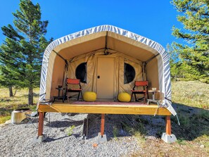 Una carpa con dos sillas y una mesa, sobre superficie de grava con árboles al fondo.
