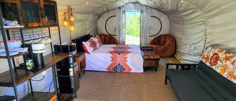 A tented accommodation with a bed, seating area, kitchenette, and a view through the entrance.