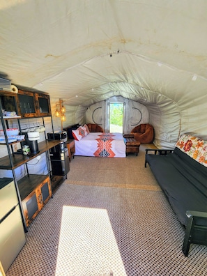 A tented accommodation with a bed, seating area, kitchenette, and a view of greenery outside.