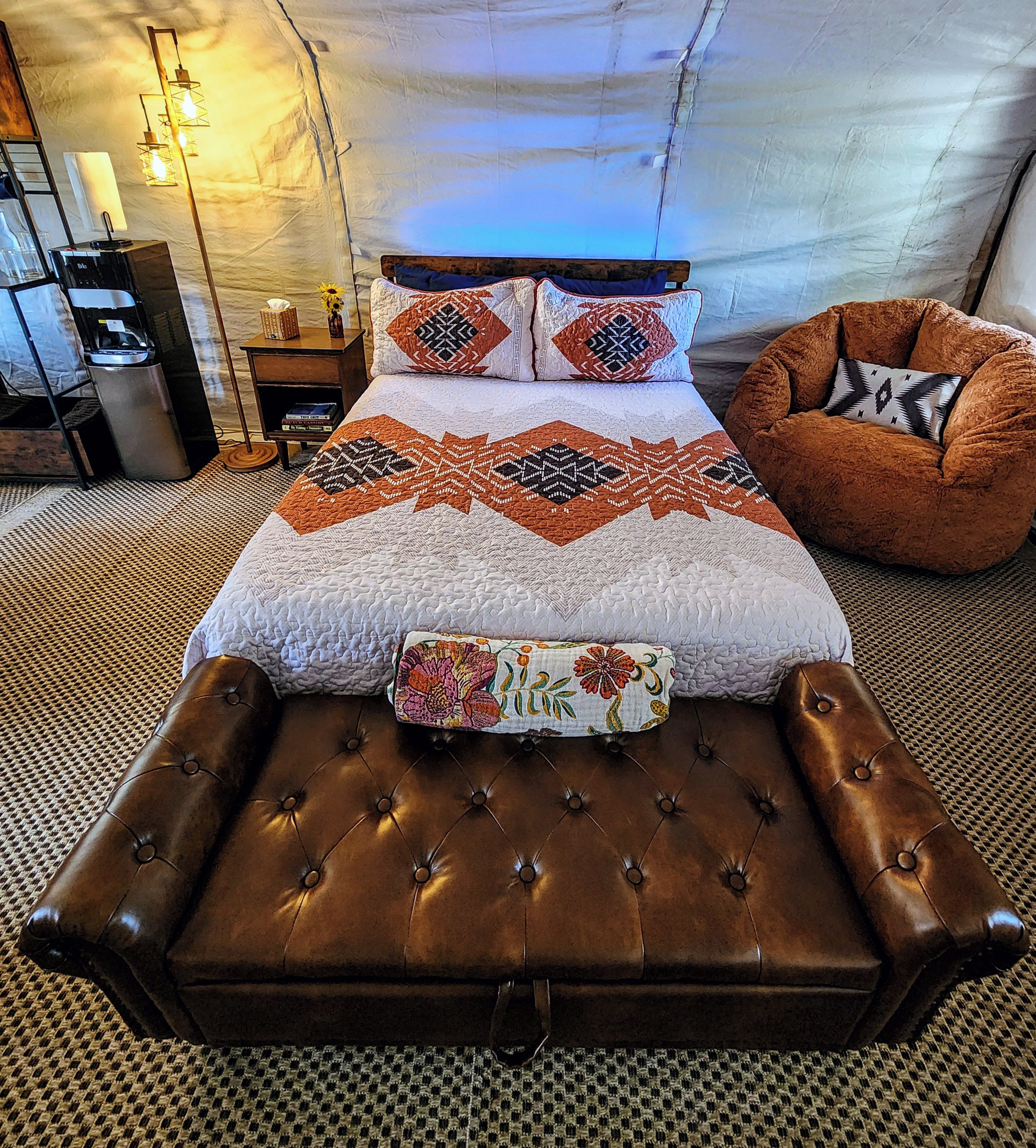 Una cama con un colcha estampada, un reposapiés de cuero marrón y una almohada decorativa.