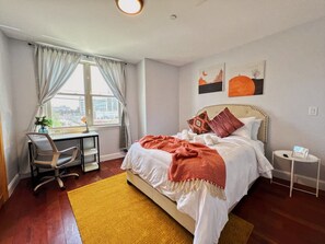 A bedroom with a bed, a desk, and a chair. There is a window with curtains, a wall with artwork, and a small table with a lamp.