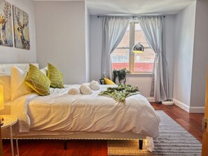 Et soveværelse med seng, puder, et natbord, en lampe, et vindue med gardiner og kunst på væggen.