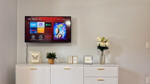 A modern living room with a flat-screen TV mounted on the wall displaying a streaming service interface. Below the TV, there is a white console with drawers and a small plant. - Ramblei near Chinatown & CenterCity: 2-Bedroom - Unit C (Philadelphia)