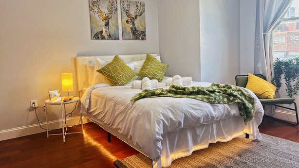 A neatly made bed with white linens and green floral throw blankets, a bedside table with a lamp, and two paintings of deer on the wall. - Ramblei near Chinatown & CenterCity: 2-Bedroom - Unit C (Philadelphia)