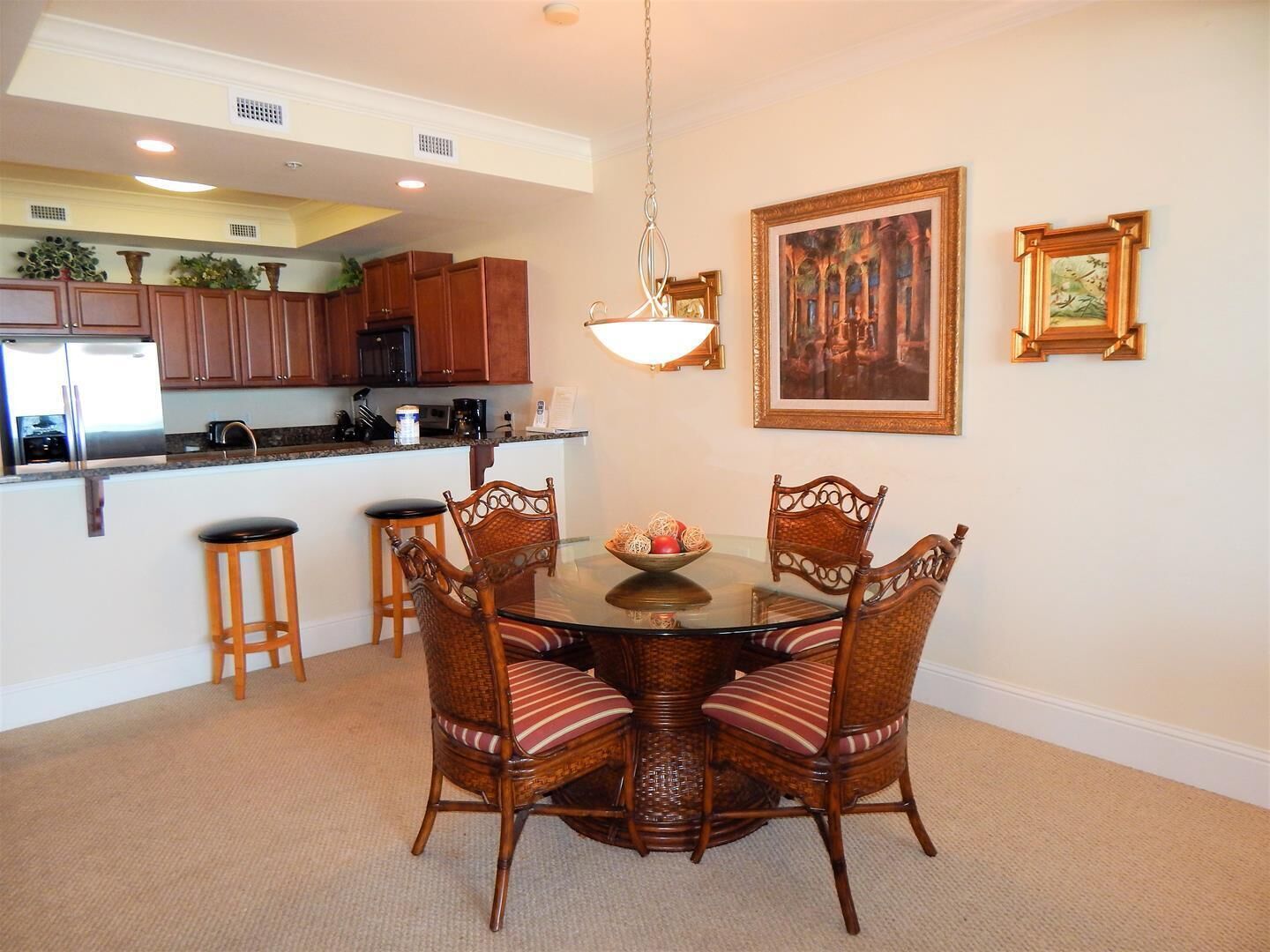 A dining area with a glass table surrounded by wicker chairs, a kitchen in the background, and two framed artworks on the wall.
