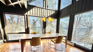 A modern dining area with a large wooden table and four chairs, large windows with a view of trees, and a hanging light fixture.