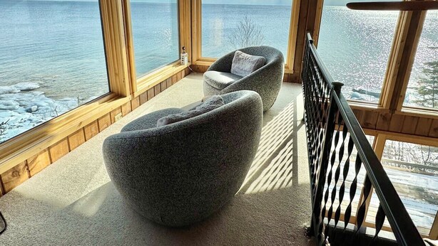 A cozy room with a large window overlooking a frozen body of water, featuring two grey upholstered chairs and a wooden floor.