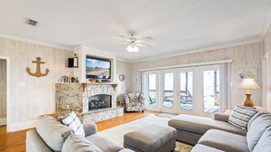 A living room with a stone fireplace, a ceiling fan, and a view through large windows. - Beautiful bayfront home with views of the Dauphin Island Bridge (Dauphin Island)