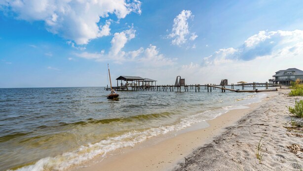 A beach with a wooden pier extending into the water, a small boat, and a building on the right. - Beautiful bayfront home with views of the Dauphin Island Bridge (Dauphin Island)