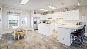 A modern kitchen with white cabinetry, a central island, and a dining area with wooden chairs. - Beautiful bayfront home with views of the Dauphin Island Bridge (Dauphin Island)