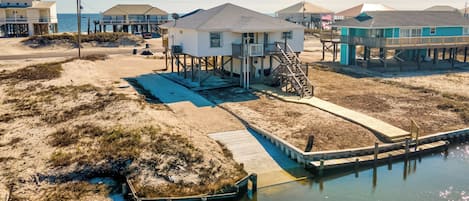 A coastal house with a dock extending into the water, surrounded by sandy terrain and other buildings.