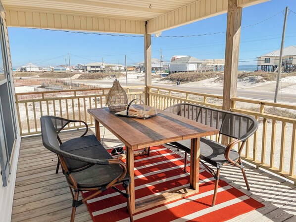 Een overdekt terras met een houten tafel en twee stoelen, een rood-wit geruit kleed en uitzicht op het strand en gebouwen in de verte.