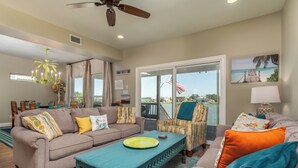 A spacious living room with a sofa, armchairs, and a coffee table. There is a balcony with a view of the water and an American flag.