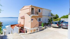 A two-story building with a balcony, a parking area with cars, and a view of the sea. - Apartments Nives (Okrug)