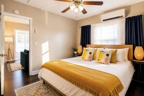 Un dormitorio con una cama, ventilador de techo, aire acondicionado, ventana con cortinas y una lámpara de noche.