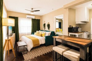 A hotel room with a bed, a desk, a chair, a small table, a lamp, a ceiling fan, an air conditioning unit, a microwave, and a kitchenette.