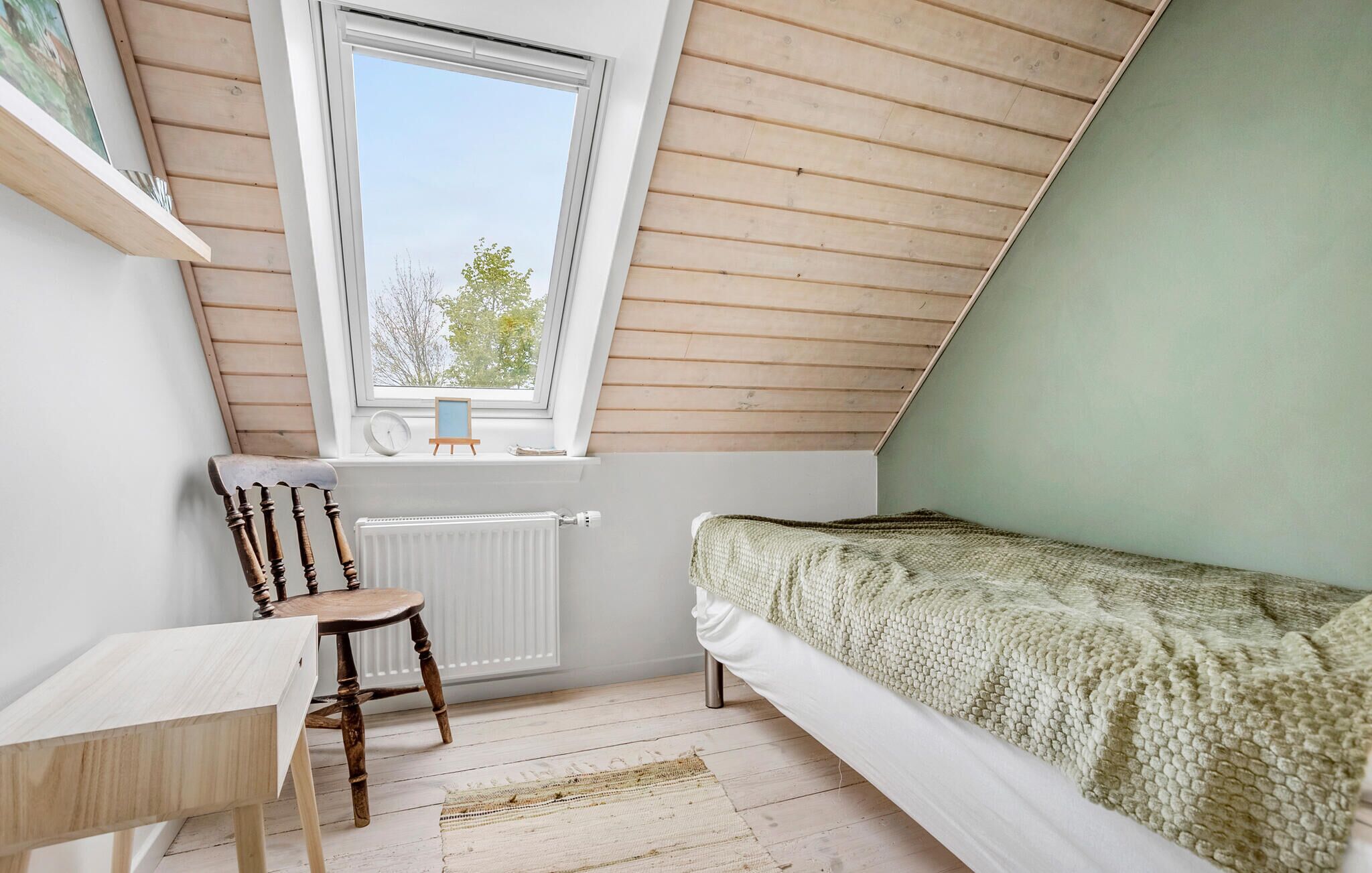 Ein kleines, gemütliches Schlafzimmer im Dachgeschoss mit schrägen Wänden, einem Holzstuhl, einem kleinen Tisch, einem Bett mit Strickdecke und einem Fenster mit Blick auf Bäume.
