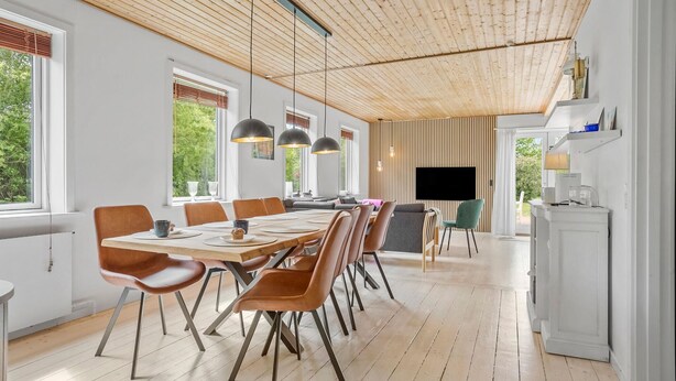 A modern dining area with wooden flooring, a long table, and multiple chairs. There are large windows providing natural light and a view of greenery outside.