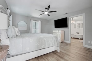 Une chambre à coucher comprenant un lit, un ventilateur de plafond, une télévision, une commode, une coiffeuse et une salle de bain avec une baignoire.