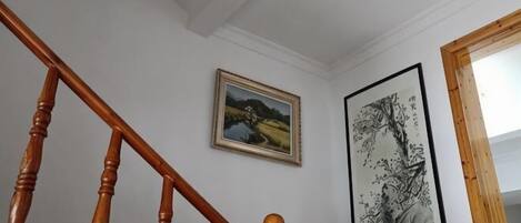 Una escalera con balaustres de madera, un cuadro con paisaje enmarco y una obra de arte vertical en la pared.