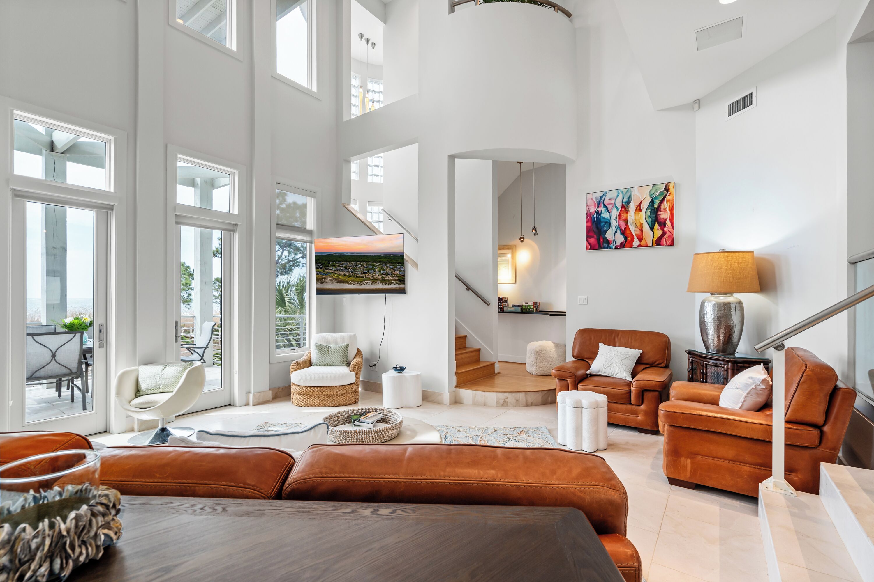 A spacious living room with a high ceiling, large windows, a staircase, and a view of the outdoors.