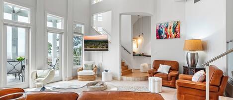 A spacious living room with a high ceiling, large windows, a staircase, and a view of the outdoors.