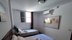 A room with two beds, a window with curtains, an air conditioning unit, and a painting on the wall.