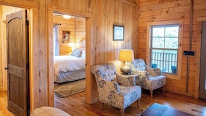 Um quarto acolhedor de cabana de madeira, com uma cama, duas poltronas estampadas, uma mesa pequena com abajur e uma janela com vista para árvores.