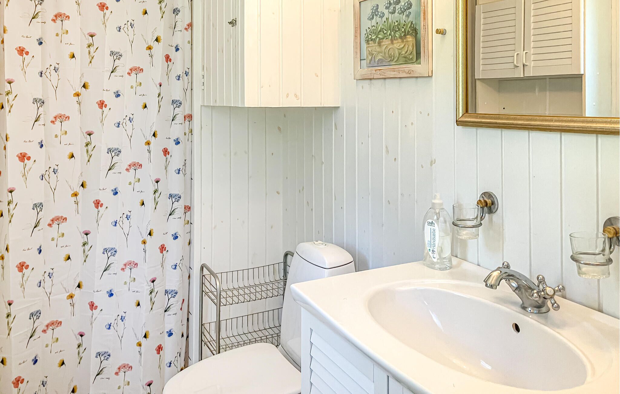 Ein Badezimmer mit einem weißen Waschbecken, einer Toilette, einem Duschvorhang mit Blumenmuster, einer kleinen Ablage für Toilettenartikel und einem gerahmten Bild an der Wand.