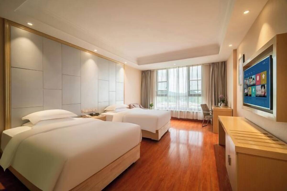 A hotel room with two beds, a desk, a TV, and large windows.